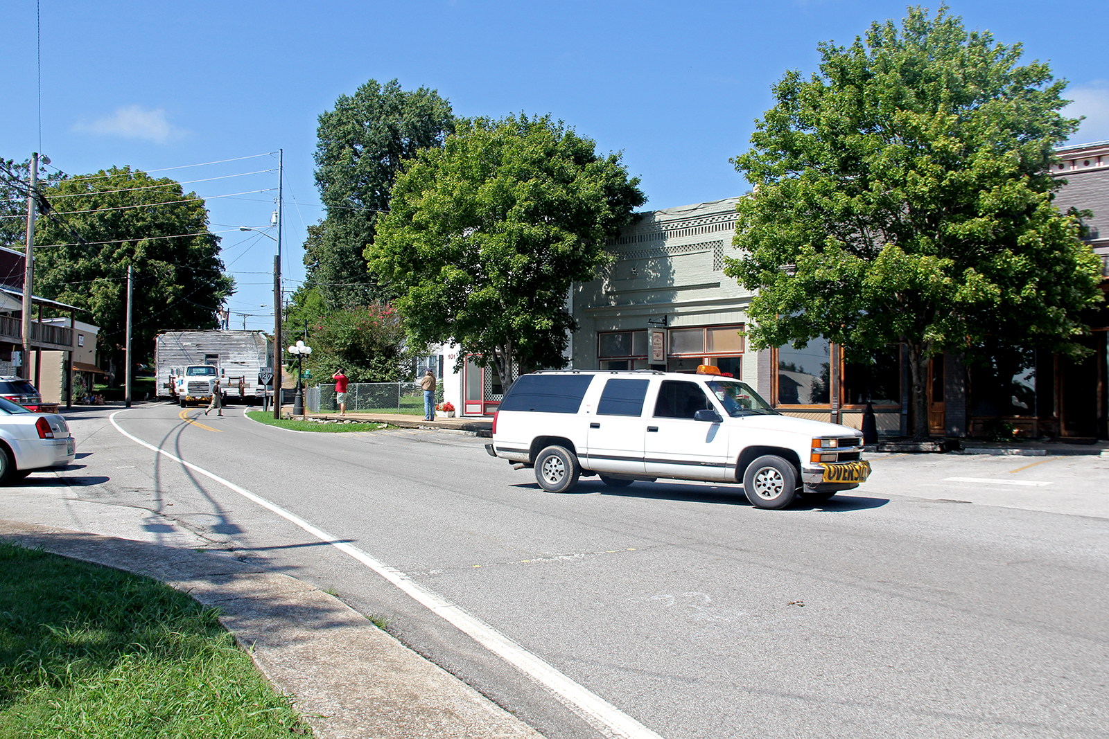 White SUV with oversized load warning driving down main street in Lynnville, TN and a truck pulling a trailer loader with the one-room school house headed for Colonel's farm