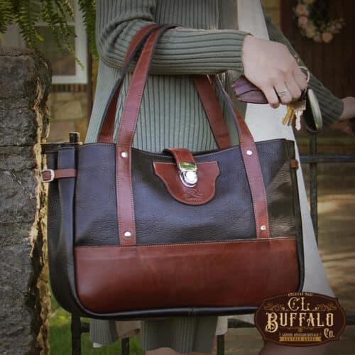 Woman carrying Bentley Tote in dark Tobacco Brown American Buffalo with Vintage Brown Steerhide trim, standing in front of iron gate in stone wall