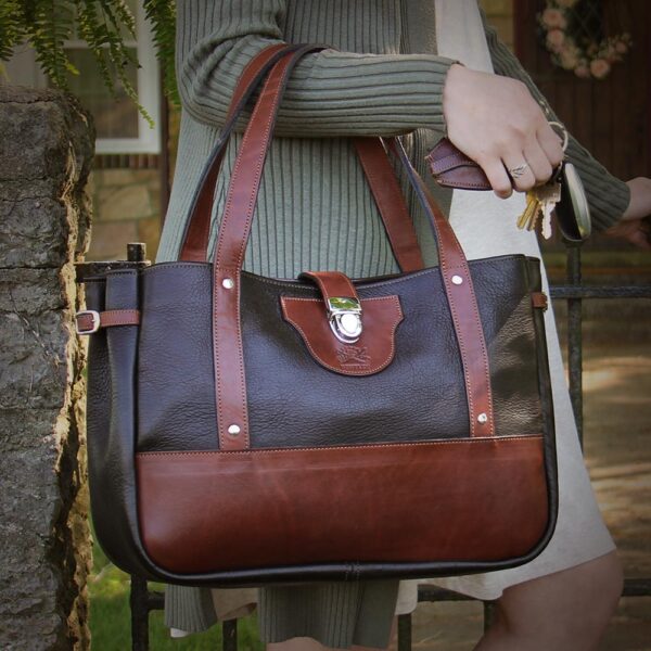 Woman carrying Bentley Tote in dark Tobacco Brown American Buffalo with Vintage Brown Steerhide trim, standing in front of iron gate in stone wall