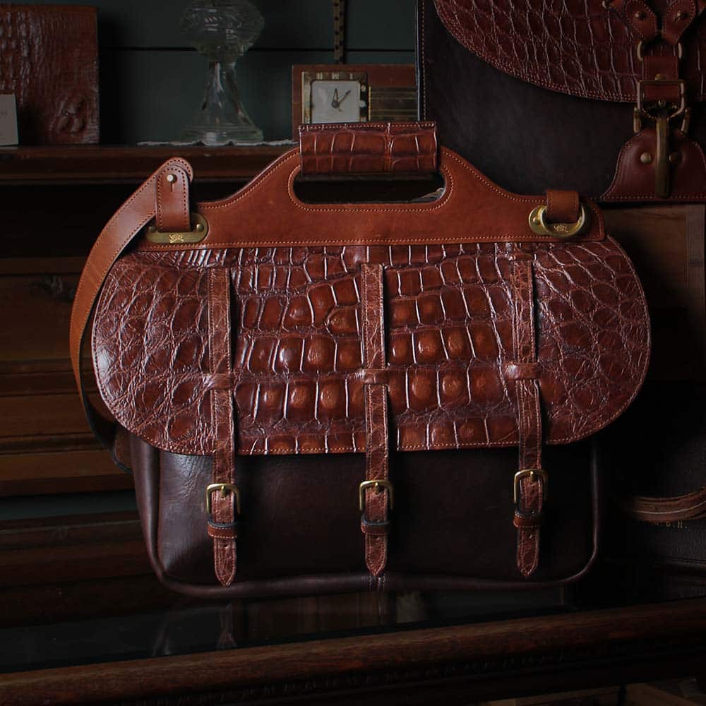 No.1 Saddlebag briefcase in Tobacco Brown American Buffalo with brown American Alligator flap and Vintage Brown Steerhide Trim resting on a glass antique display cabinet