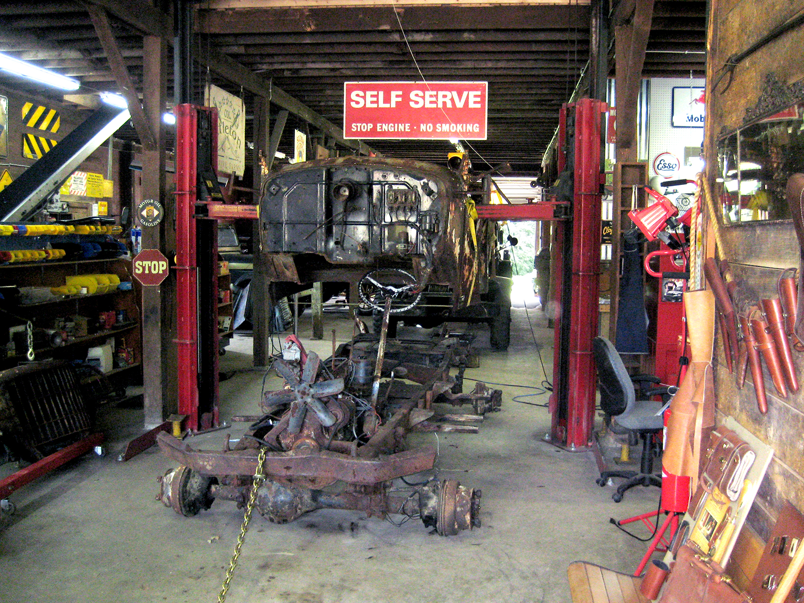 Very rusted chassis of 1941 Army Command Car on floor of Colonel's Hot Rod Garage with the rusted body suspended on lift over it.