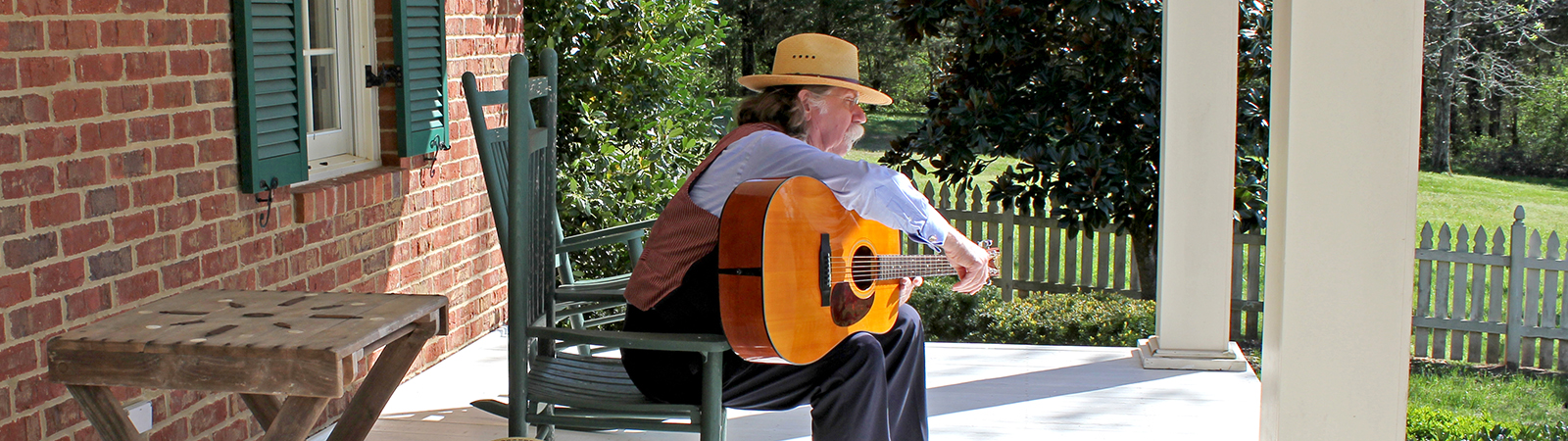 Colonel sitting in rocking chair on front porch playing guitar.