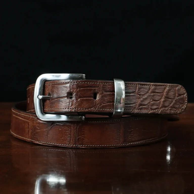 An American Alligator Belt with Nickel hardware showing the front coiled view on a wooden table with black background - id 001