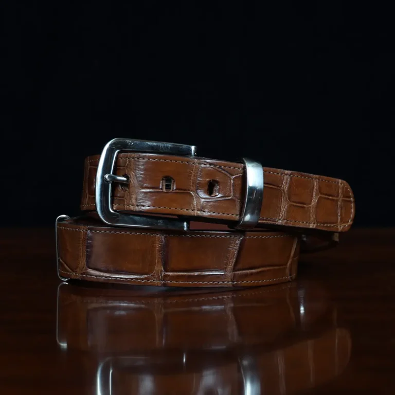 An American Alligator Belt with Nickel hardware showing the coiled front view on a wooden table with black background - id 001