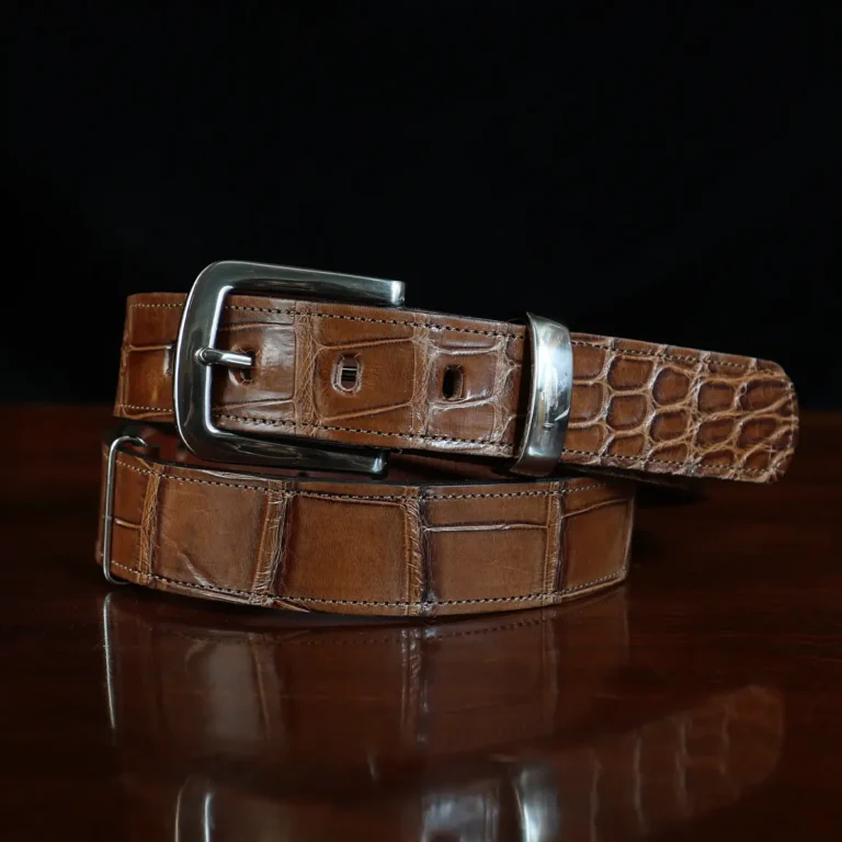 An American Alligator Belt with Nickel hardware showing the front coiled view on a wooden table with black background