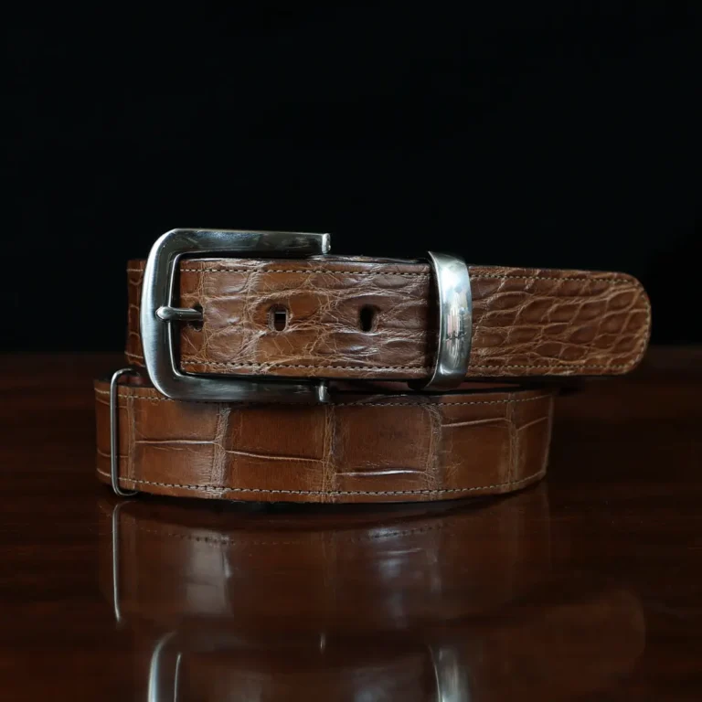 An American Alligator Belt with Nickel hardware showing the coiled front view on a wooden table with black background - id 001