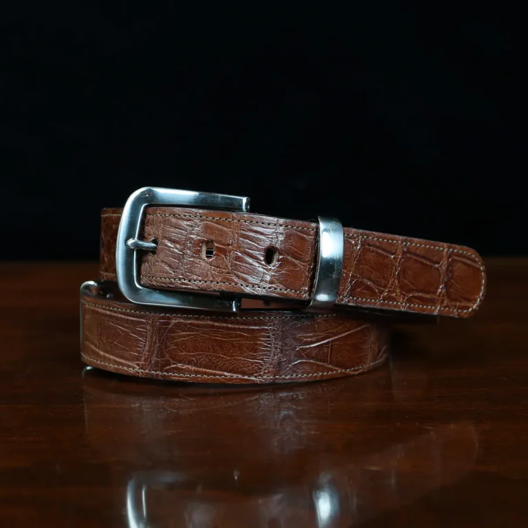 An American Alligator Belt with Nickel hardware showing the coiled front view on a wooden table with black background - id 001