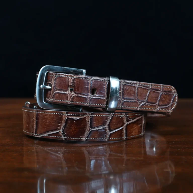 An American Alligator Belt with Nickel hardware showing the coiled front view on a wooden table with black background - id 001