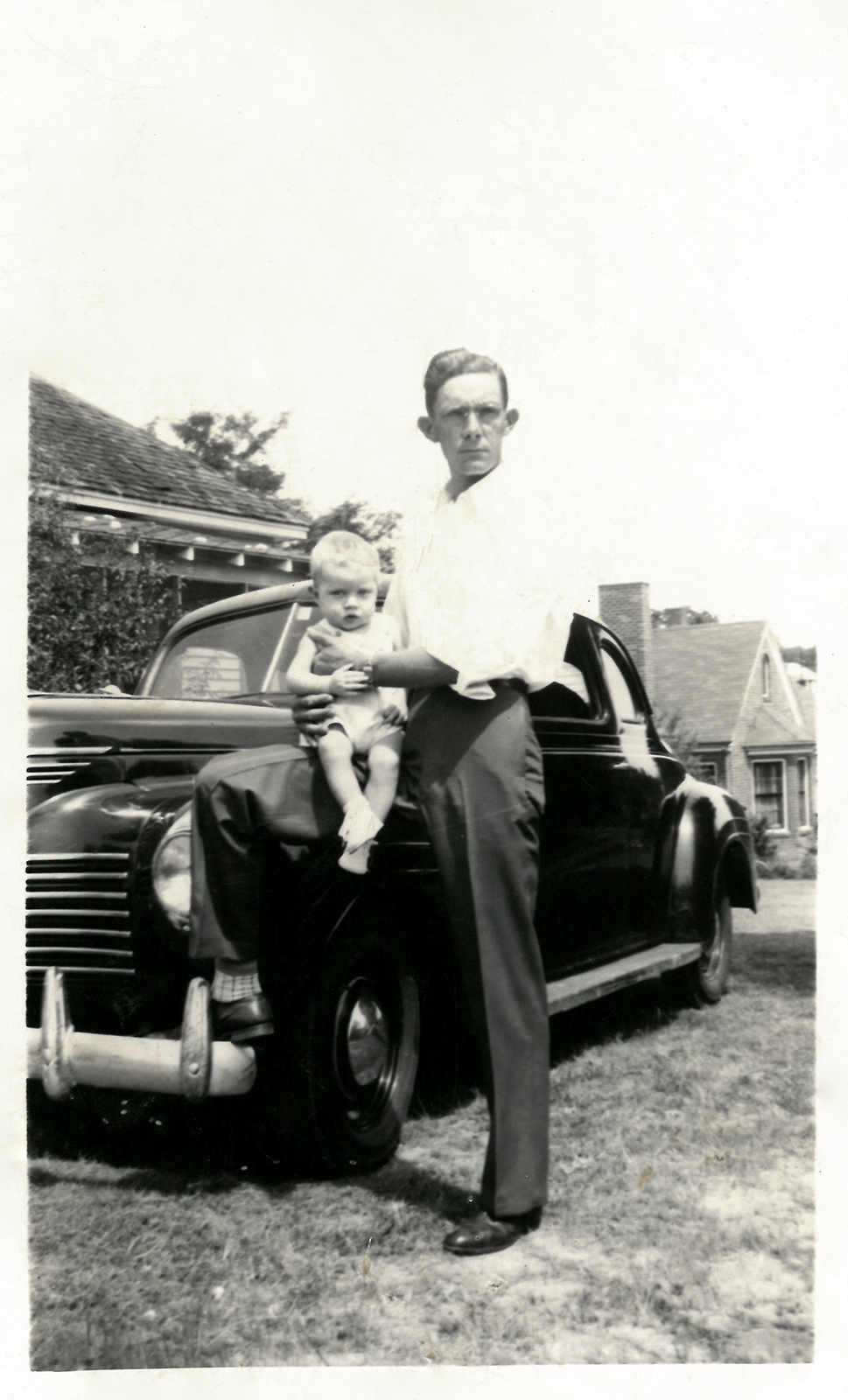 Black and white photo of Colonel's dad, called Pap, holding Colonel as a baby on his knee while standing next to the hood of an old car