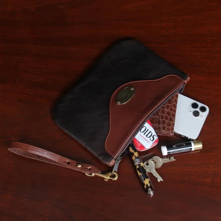 open view brown leather wristlet clutch on black background and wood table with items spilling out