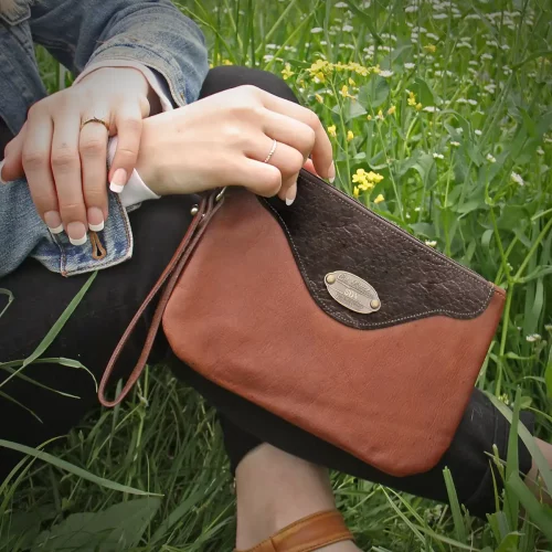 closeup of model holding brown leather wristlet clutch in lifestyle setting of green grass