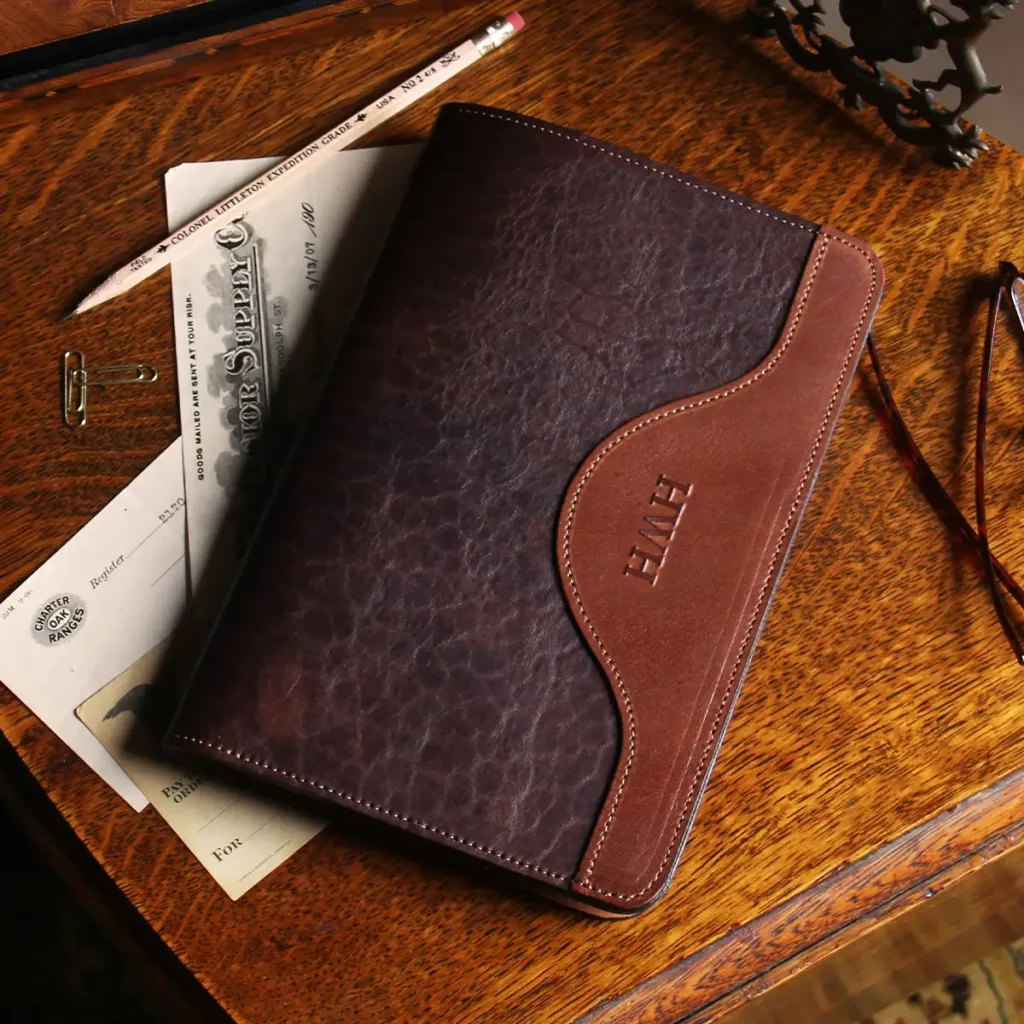 Leather travel portfolio in tabacco brown american buffalo on a table with paper, glasses, pencil, and paper