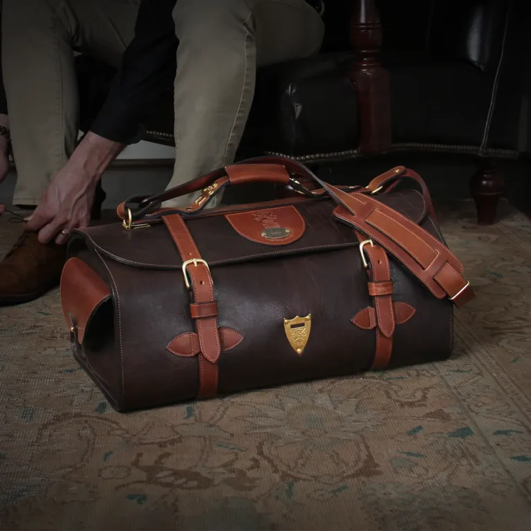 No. 2 Duffel Bag in Tobacco Brown American Buffalo with Vintage Brown American Steerhide trim - Front view of back resting on the floor next to a man tying his shoe in an antique chair