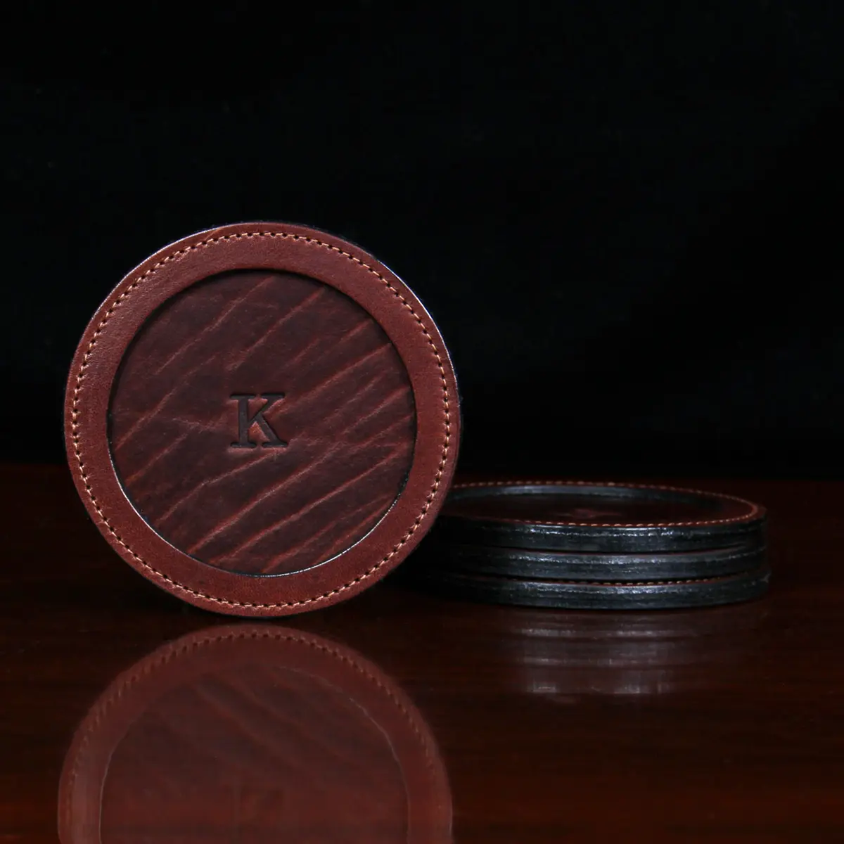 leather coaster with K initial on table sitting next to 3 others stacked next to it