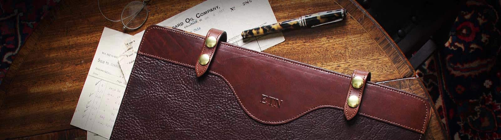 Portfolios and Journals Category header image: Close cropped image of the No. 28L Legal portfolio in dark Tobacco Brown American Buffalo with vintage brown steerhide trim on a desk with a nice pen and a few papers