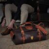 Dark brown buffalo leather No. 2 Duffel bag on floor in front of man tying shoes in a leather chair with dramatic lighting