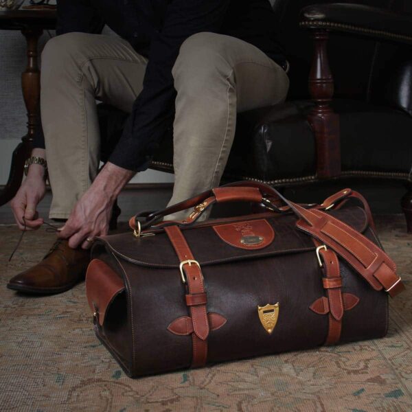 Dark brown buffalo leather No. 2 Duffel bag on floor in front of man tying shoes in a leather chair with dramatic lighting
