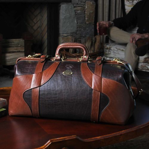 Vintage-style Gladstone dark brown american buffalo leather No. 5 Grip travel bag on wooden coffee table with a stone fireplace and a man sitting in a wingback chair in the background