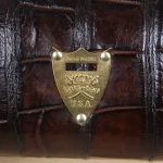 close-up view of pommenl sheild howing serial number on brown american alligator duffel bag on cream background