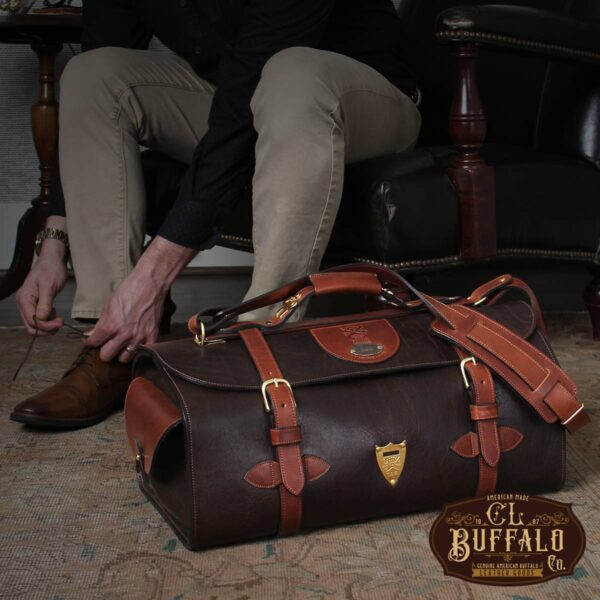 No. 2 Duffel Bag in Tobacco Brown American Buffalo with Vintage Brown American Steerhide trim - Front view of back resting on the floor next to a man tying his shoe in an antique chair