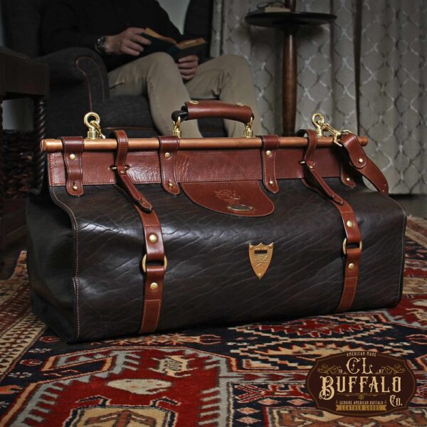 Dark brown buffalo leather No. 3 grip travel bag sitting on an antique carpet with a man reading in a chair in the background