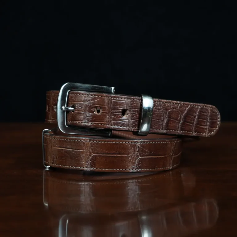 An American Alligator Belt with Nickel hardware showing the front coiled view on a wooden table with black background - id 002
