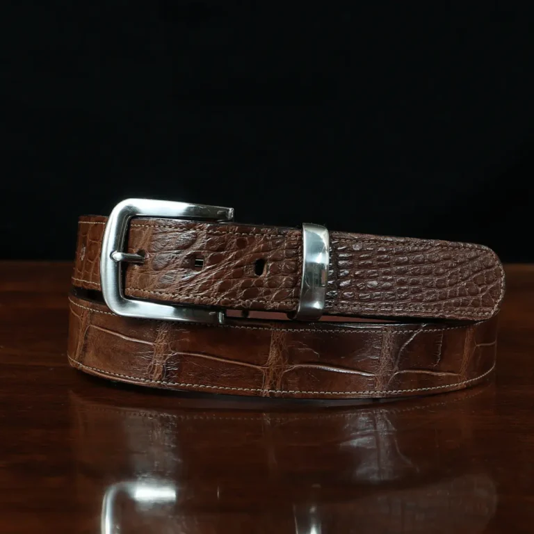 An American Alligator Belt with Nickel hardware showing the front coiled view on a wooden table with black background - id 002