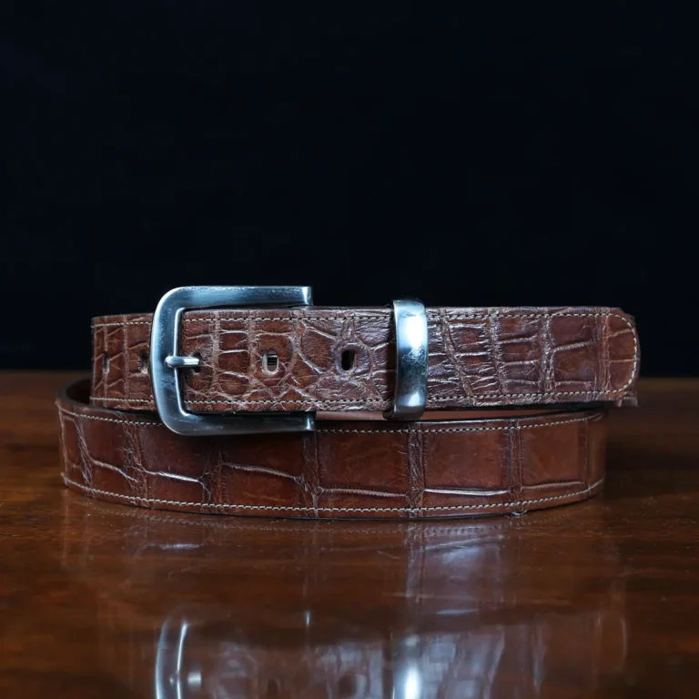 An American Alligator Belt with Nickel hardware showing the front coiled view on a wooden table with black background - id 002